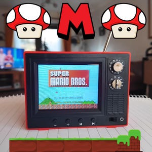 Puede incluir: Un televisor retro rojo y negro con una pantalla que muestra el título "SUPER MARIO BROS.". El televisor tiene una pequeña antena y una perilla de volumen. El televisor está sentado sobre una superficie blanca y gris.