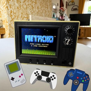 Op de afbeelding: Een miniatuurtelevisie in retrostijl met het spel "Metroid". Het scherm is zwart met een gele lijst, en de titel van het spel is zichtbaar. Extra gamecontrollers zijn rond de tv geplaatst, waaronder een wit-zwarte controller, een blauwe controller en een handheld console.