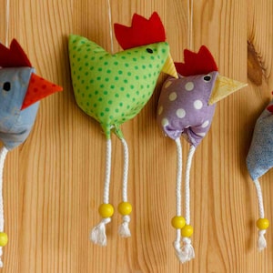 Peut inclure: Quatre poulets en tissu colorés avec des crêtes et des becs rouges suspendus à un mur en bois. Chaque poulet a des pattes en ficelle blanche avec des perles jaunes.