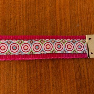 Könnte beinhalten: Ein rosa-weißer Schlüsselanhänger mit einem silbernen Schlüsselring. Der Schlüsselanhänger hat einen gemusterten Stoff mit Kreisen in Grün-, Blau- und Rottönen.