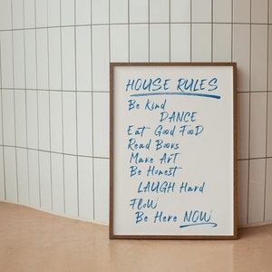 May include: A framed white board with the words "HOUSE RULES" written in black marker. The board lists a set of rules written in blue marker, including "Be Kind", "Dance", "Eat Good Food", "Read Books", "Make Art", "Be Honest", "Laugh Hard", "Flow", and "Be Here Now".