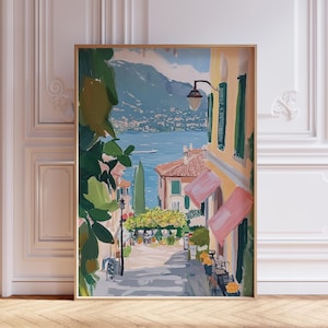 May include: A painting of a colorful Italian village with a view of a lake and mountains in the background. The painting is framed in a gold frame and is hanging on a white wall.