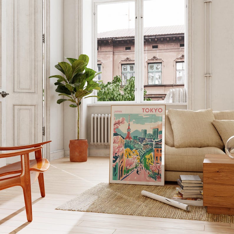 May include: A colorful poster with the text "TOKYO" and "JAPAN" in a white frame. The poster depicts a cityscape with a tower and cherry blossoms. The poster is leaning against a white wall in a room with a wooden floor, a large plant, and a couch.