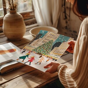 May include: A hand holds a colorful illustration of a snowy city scene with a large Christmas tree in the center. People are ice skating on a frozen pond in front of the tree. The illustration is on a white background.