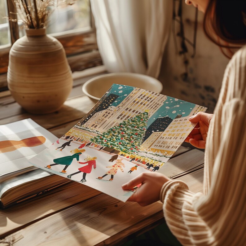 May include: A hand holds a colorful illustration of a snowy city scene with a large Christmas tree in the center. People are ice skating on a frozen pond in front of the tree. The illustration is on a white background.