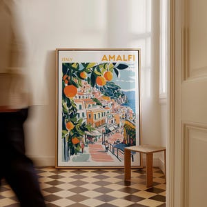 May include: A framed print of a colorful illustration of the Amalfi Coast in Italy. The print features a cityscape with buildings, a winding road, and a lush orange tree. The text "ITALY" and "AMALFI" is printed in the top left and right corners of the print.