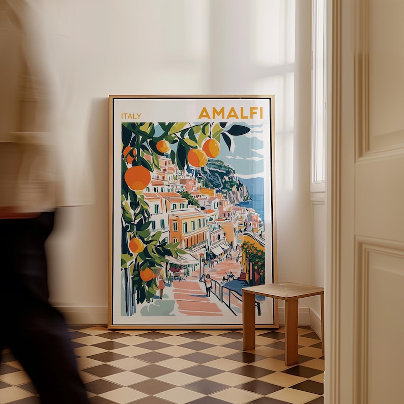 May include: A framed print of a colorful illustration of the Amalfi Coast in Italy. The print features a cityscape with buildings, a winding road, and a lush orange tree. The text "ITALY" and "AMALFI" is printed in the top left and right corners of the print.