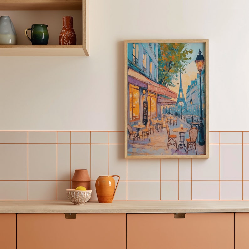 May include: A framed painting of a Parisian street scene with a cafe, tables, chairs, and the Eiffel Tower in the distance. The painting is in a light wood frame and is hanging on a white tiled wall.