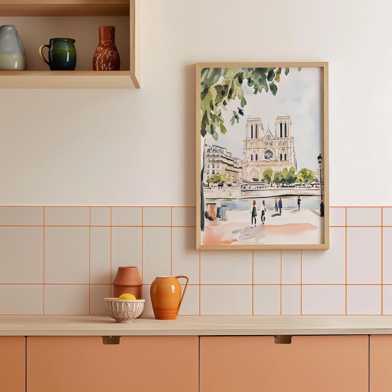 May include: A watercolor painting of the Notre Dame Cathedral in Paris, France. The painting is framed in a light wood frame and hangs on a white tiled wall in a kitchen.