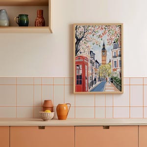 May include: A framed illustration of a street scene in London, England. The illustration features a red telephone booth, a clock tower, and a row of buildings with blooming trees. The illustration is framed in a light wood frame.