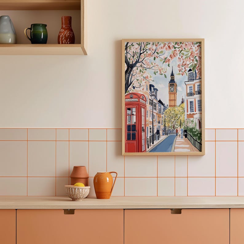 May include: A framed illustration of a street scene in London, England. The illustration features a red telephone booth, a clock tower, and a row of buildings with blooming trees. The illustration is framed in a light wood frame.