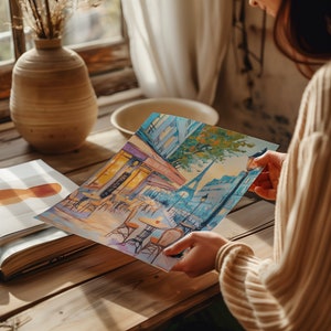 May include: A person is holding a painting of a Parisian street scene with a cafe and the Eiffel Tower in the background. The painting is done in a colorful, impressionistic style.