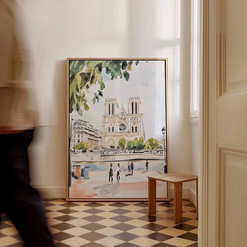 May include: A watercolor painting of the Notre Dame Cathedral in Paris, France. The painting is framed in a gold frame and is hanging on a white wall. The painting shows the cathedral from a distance, with the Seine River in the foreground. There are people walking along the riverbank.