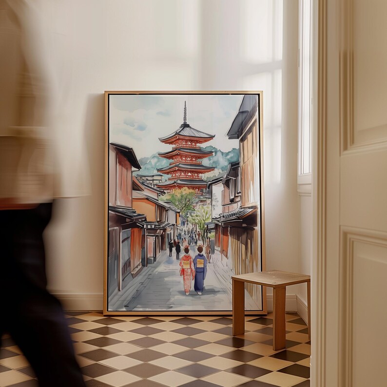 May include: Watercolor painting of a Japanese street scene with a pagoda and traditional buildings. Two figures in kimonos walk down the street. The artwork is framed in a gold-colored frame and leans against a white wall.