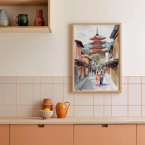 May include: Watercolor print of a Japanese street scene with a red pagoda in the background. The artwork depicts a street lined with traditional buildings, with two figures in kimonos walking away from the viewer. The print is framed in a light wood frame.