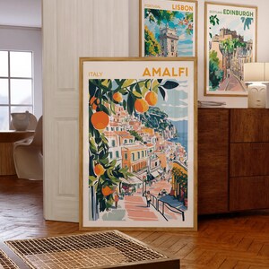 May include: Three framed travel posters with colorful illustrations of Amalfi, Lisbon, and Edinburgh. The Amalfi poster features a vibrant orange tree with fruit and a view of the city.