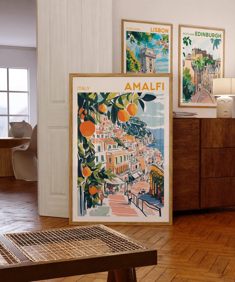 May include: Three framed travel posters with colorful illustrations of Amalfi, Lisbon, and Edinburgh. The Amalfi poster features a vibrant orange tree with fruit and a view of the city.