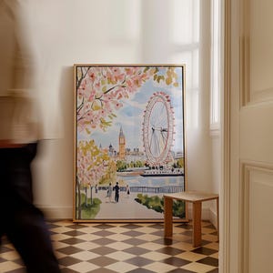 May include: A watercolor painting of the London Eye Ferris wheel with pink cherry blossoms in the foreground. The painting is framed in a light wood frame and is hanging on a white wall.