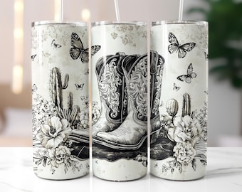 May include: Two black and white cowboy boots with floral and cactus designs on a white background. The boots are on a white tumbler with a straw.