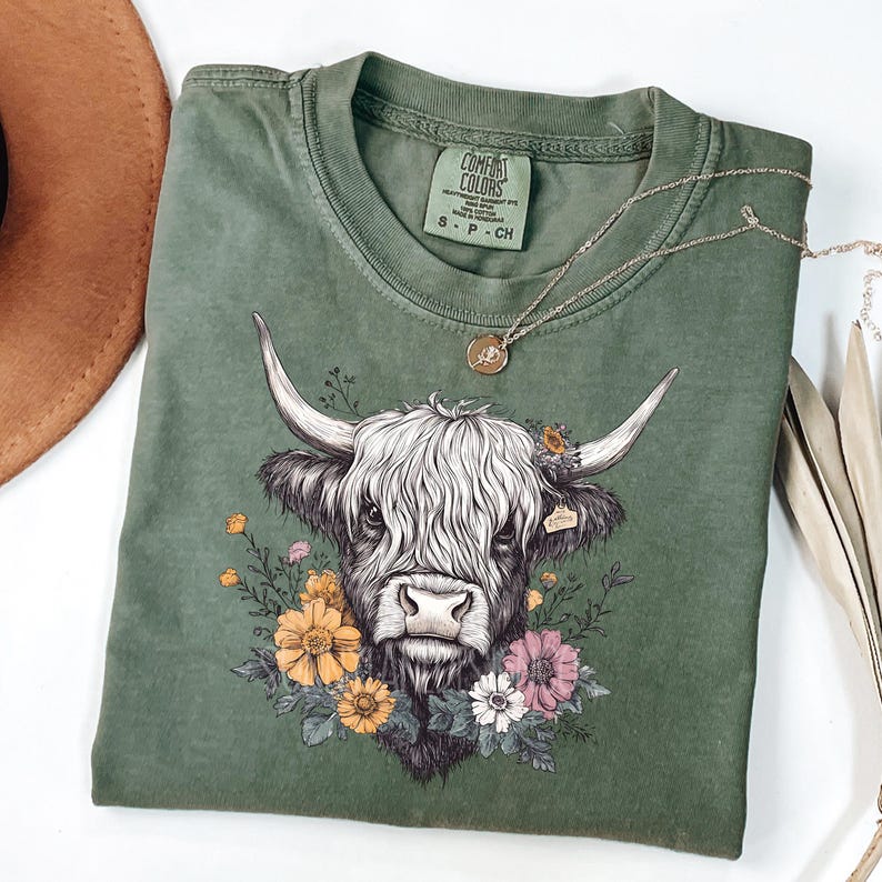 May include: A green t-shirt with a white highland cow illustration wearing a flower crown. The cow has a black nose and hooves. The shirt has a tag that says "Comfort Colors" on it.