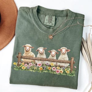 May include: A green t-shirt with a graphic of four white sheep sitting in a row on a wooden fence. The sheep are surrounded by colorful flowers. The t-shirt has a tag that says "Comfort Colors" and "S - P - CH".