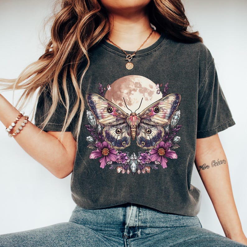 May include: Dark grey t-shirt featuring a large butterfly design with a full moon backdrop. The butterfly has purple and pink wings, surrounded by purple flowers and crystals. The shirt is a casual, comfortable style.