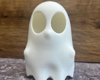 May include: A white, 3D-printed ghost figurine with large, oval eye sockets. The ghost has a rounded body and a wavy bottom edge. The background is a wooden surface.