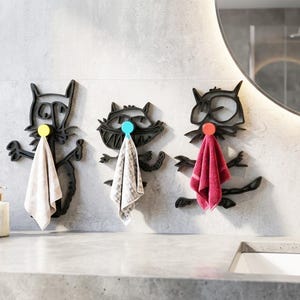 Puede incluir: Tres perchas negras con forma de mapache con pomos de colores, cada una sosteniendo una toalla de diferente color. Las perchas están montadas en una pared gris claro en un baño. Una pila de toallas blancas y un dispensador de jabón están en el mostrador.