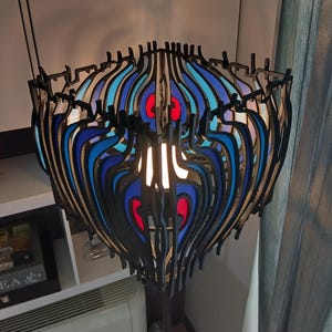 Puede incluir: Lámpara colgante moderna con un diseño en capas. La lámpara tiene un marco negro con detalles en azul, rojo y blanco. La luz está encendida, iluminando el interior. El diseño general es artístico y decorativo.