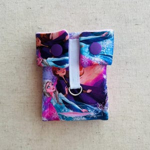 Könnte beinhalten: Eine lila und blaue Stofftasche mit einem weißen Band und zwei lila Knöpfen. Die Tasche zeigt einen Aufdruck mit Figuren aus dem Film Frozen.