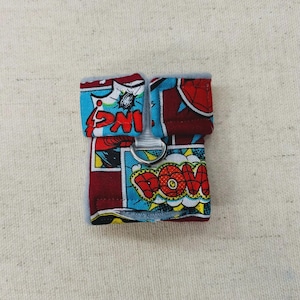 Könnte beinhalten: Kleines, quadratisches Haustierzubehör mit Comic-Thema. Das Zubehör ist hauptsächlich rot und blau, mit dem Wort "POW" in einer Sprechblase. Es hat einen silbernen Metallring und ein graues, flauschiges Futter.
