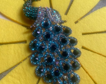 May include: A jeweled peacock brooch with a teal and dark blue color scheme. The brooch is set against a yellow background. The peacock's tail feathers are a prominent feature, with large, round, dark blue gems.