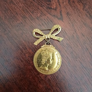 Op de afbeelding: Goudkleurige broche met een strikvormig ontwerp aan de bovenkant en een ronde hanger. De hanger toont een profiel van een gekroonde figuur en de inscriptie "QUEEN ELIZABETH II".