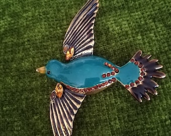 以下が含まれることがあります： 金と黒のアクセントが付いたターコイズブルーのエナメル製の鳥のブローチ。鳥は長く尖ったくちばしを持ち、羽を広げています。鳥は小さな透明なラインストーンで飾られています。