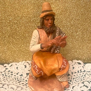 Può includere: Una statuetta in ceramica di una persona che indossa un cappello marrone e un abito con motivi gialli e marroni. La persona tiene un piccolo oggetto tra le mani.