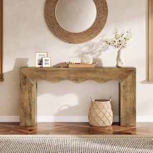 May include: A light brown wooden console table with a scalloped apron design. On the table are two framed pictures, a rectangular woven tray holding a white decorative sphere, and a white vase with white flowers. A woven basket sits below.