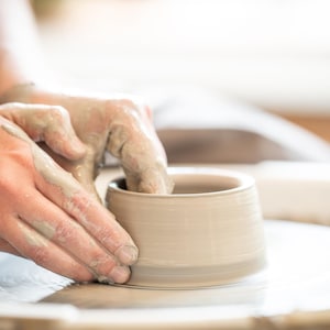 May include: Hands covered in wet clay moulding a small, light-coloured ceramic bowl on a pottery wheel. The wheel and bowl are white. The scene is well-lit, with a soft focus on the hands and the clay vessel.