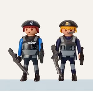 Op de afbeelding: Twee speelgoedfiguren van politieagenten. Elke figuur draagt een zwarte baret, een grijs en zwart vest en houdt een zwart wapen vast. De ene figuur heeft rood haar en een blauw shirt, de andere heeft blond haar en een marineblauw shirt.