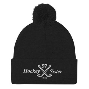 Puede incluir: Gorro de punto negro con un pompón blanco en la parte superior. El gorro tiene un diseño bordado en blanco de dos palos de hockey cruzados con un disco y el número 97. El texto "Hockey Sister" está bordado debajo de los palos.