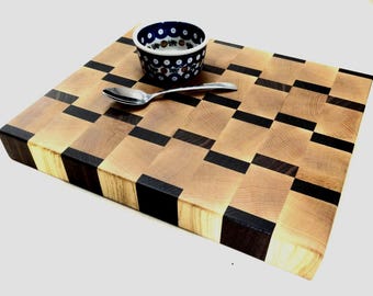 May include: A wooden cutting board with a checkerboard pattern of light and dark wood. A small blue and white bowl and a silver spoon rest on the board. The board is approximately 12 inches square.