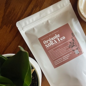 Peut inclure: Un sachet blanc de "Organic NORA Tea" est posé sur une surface en bois, à côté d'une plante en pot. L'étiquette est de couleur rose poussiéreux avec le texte "Beautiful One Midwifery". Le thé est un thé nourrissant traditionnel pour la fertilité, la grossesse et la récupération post-partum.