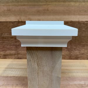May include: A white post cap with a decorative design sits atop a natural wood post. The post cap has a layered, architectural style. The wood post has visible grain and is square in shape. The background is a wooden surface.