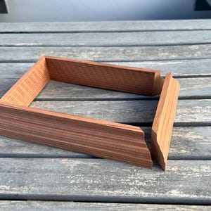 May include: Two pieces of unfinished wood frame, made of cedar, are laid out on a gray wooden surface. The frame is rectangular and has a simple design.