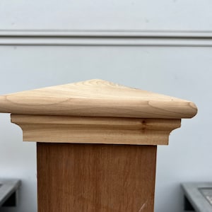May include: A wooden fence post topper with a triangular top and a decorative edge. The topper is made of natural wood and has a light brown color.