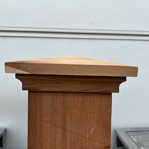 May include: A wooden fence post with a triangular cap. The cap has a decorative molding around the edge.