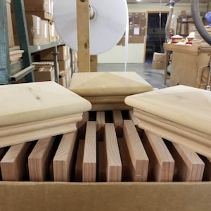May include: Three unfinished wooden square post caps with a pyramid-shaped top. The caps are stacked on top of each other and are sitting on a pile of wooden slats.