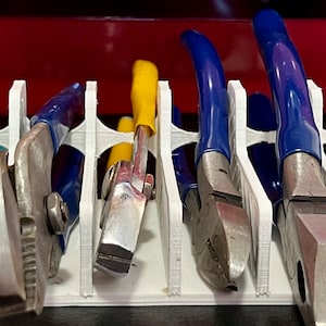 Peut inclure: Un organisateur d'outils blanc imprimé en 3D contenant divers outils à main, notamment des pinces et des tournevis. Les outils ont des poignées bleues, jaunes et noires. L'organisateur est dans un tiroir de boîte à outils rouge.