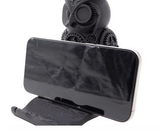 Puede incluir: Un soporte para teléfono negro con forma de búho con un diseño steampunk, que sostiene un teléfono inteligente negro. El búho tiene intrincados detalles de engranajes y un monóculo. El teléfono está inclinado para su visualización.