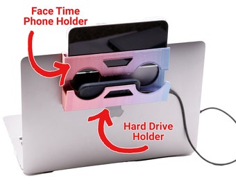 Puede incluir: Un soporte para teléfono y disco duro impreso en 3D de color rosa y morado, fijado a la parte posterior de un portátil plateado. El soporte tiene dos ranuras, una para un teléfono y otra para un disco duro. El texto "Face Time Phone Holder" está escrito encima de la ranura para el teléfono y "Hard Drive Holder" está escrito debajo de la ranura para el disco duro.