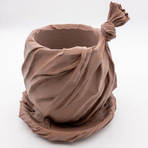May include: A brown ceramic planter shaped like a tied bag. The planter has a round opening and sits on a matching saucer base. The bag-like form features detailed folds and a knotted top.